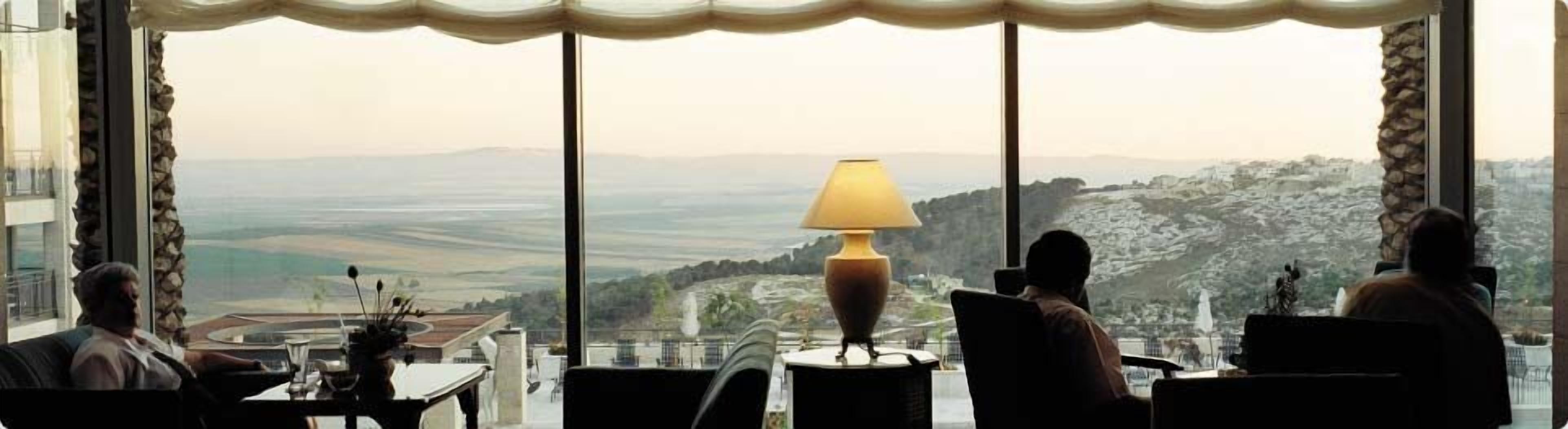 A room with a large window offering a view of a coastal landscape. There is a table with a lamp and a chair.