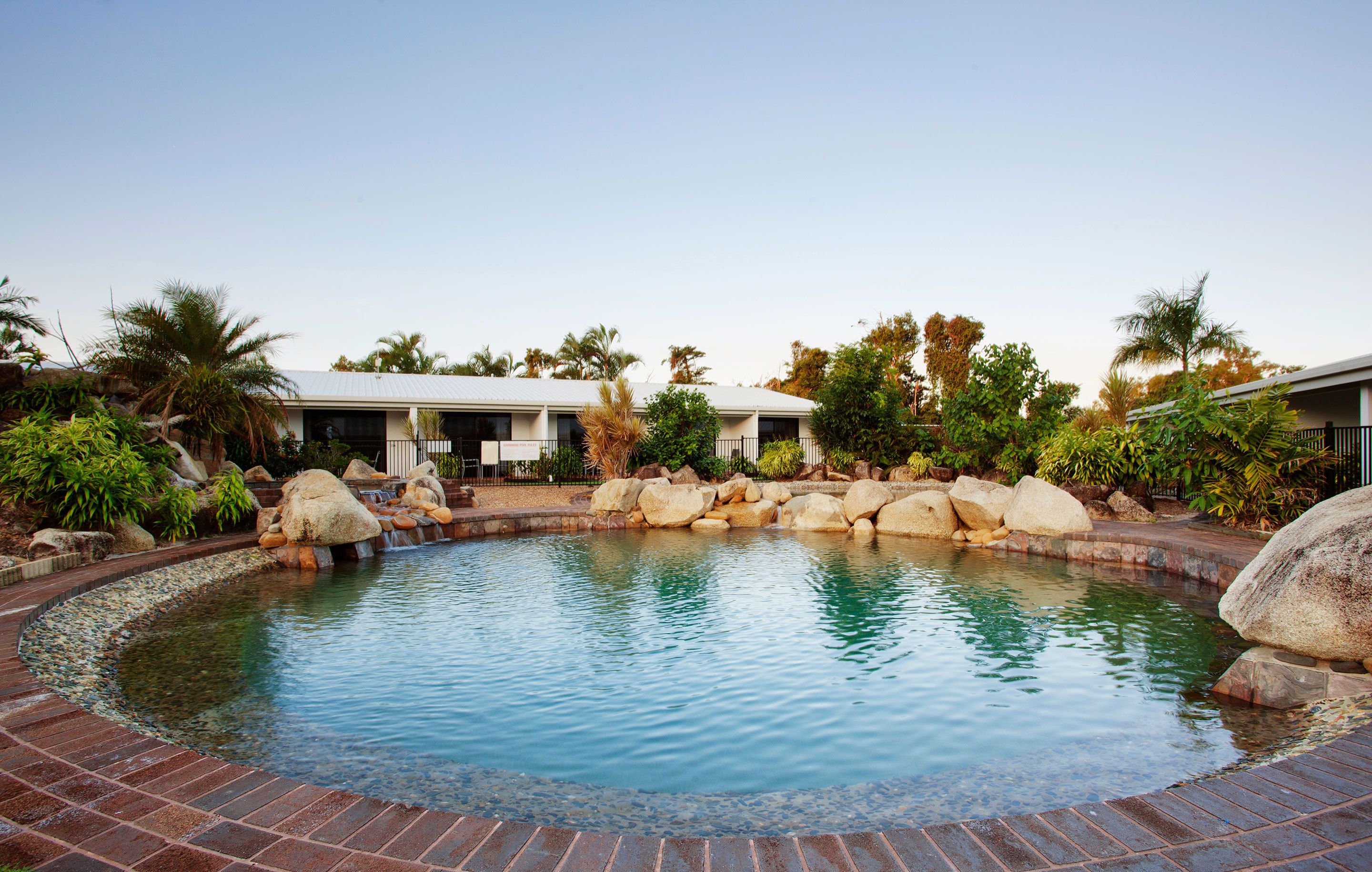 Una piscina con borde de ladrillo, rodeada de rocas y palmeras.