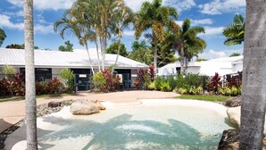 Un área de piscina con agua cristalina, rodeada de palmeras y plantas tropicales, con un edificio al fondo.