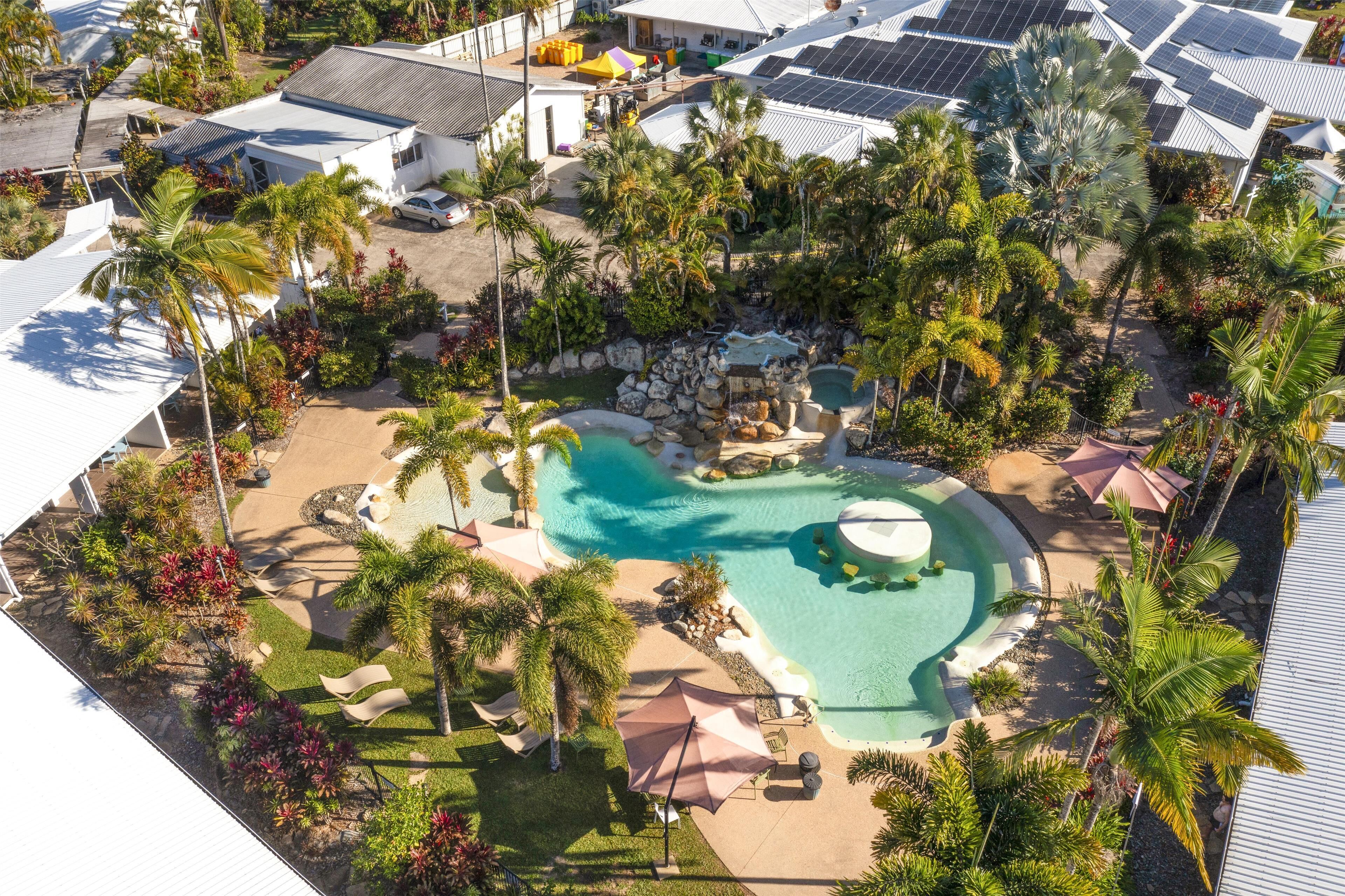 An aerial view of a resort with a central swimming pool, surrounded by palm trees and a rocky waterfall feature.
