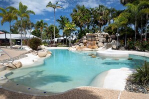 Una piscina cristalina con una cascada, rodeada de palmeras y un muro de piedra.
