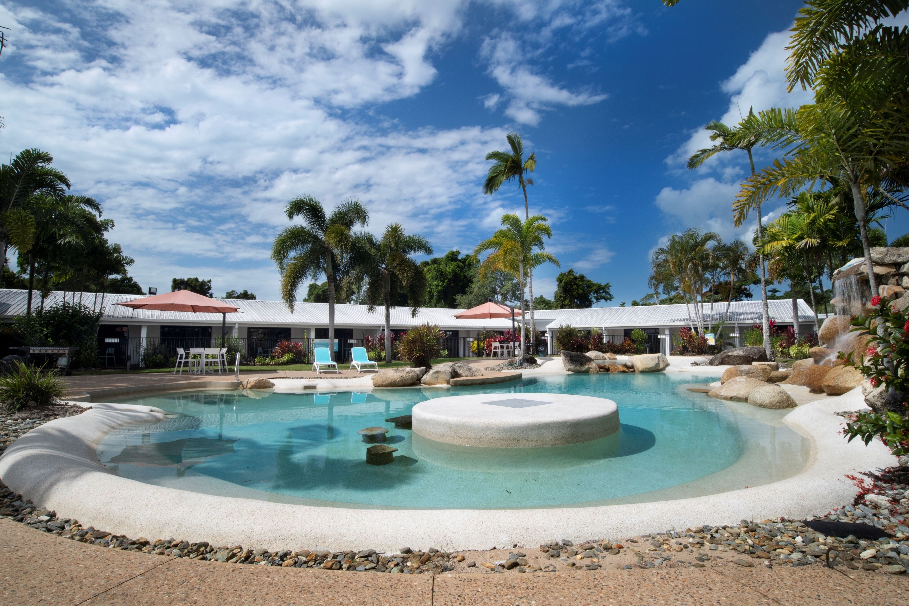 A swimming pool with a circular hot tub, surrounded by palm trees and rocks.