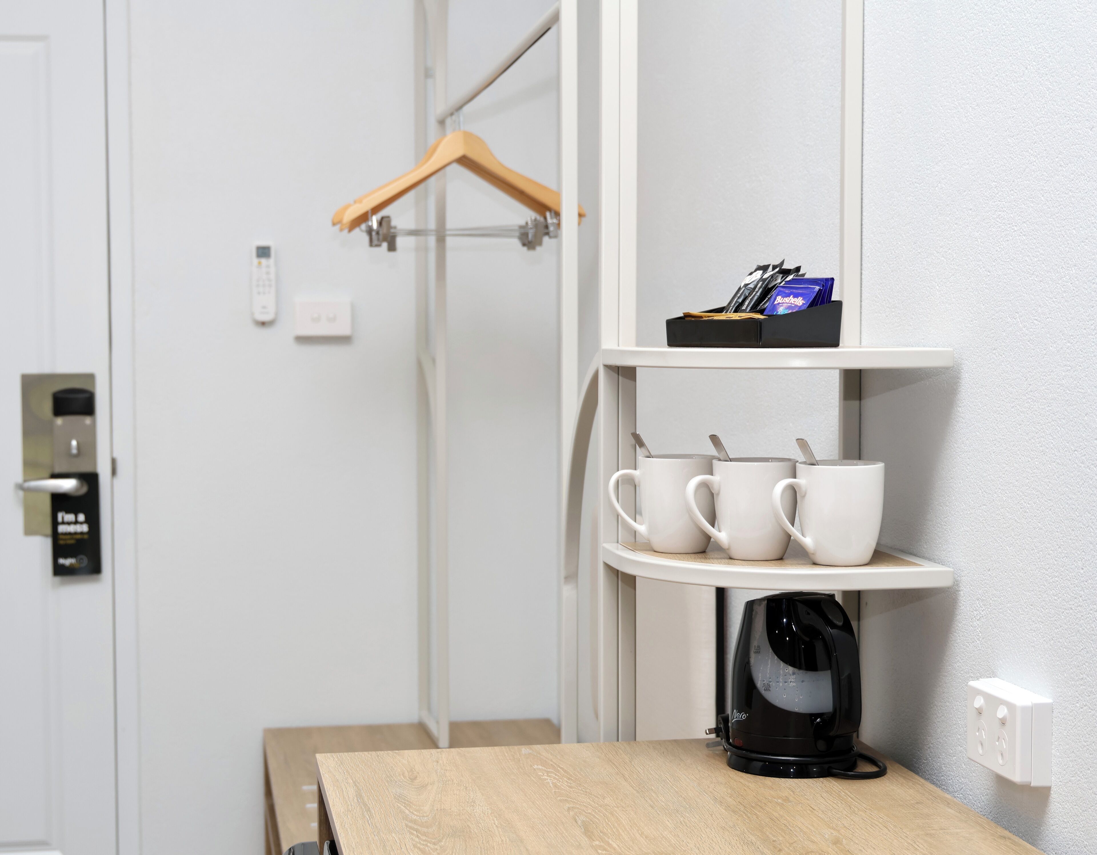 Ein Hotelzimmer mit Holztisch, einer schwarzen Kaffeemaschine, weißen Tassen, einem Kleiderständer und einem Regal mit Pflegeprodukten.