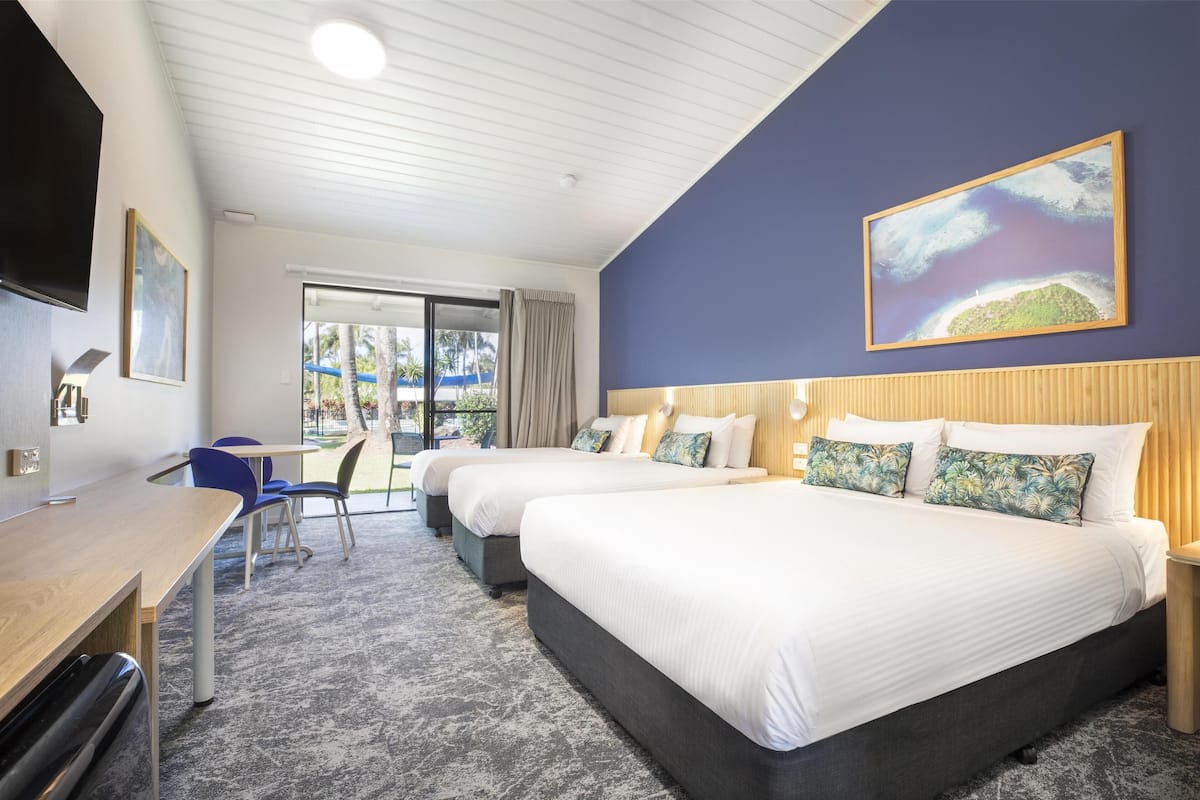 A hotel room with two beds, a desk, and a chair. There is a large window with a view of palm trees and a beach.