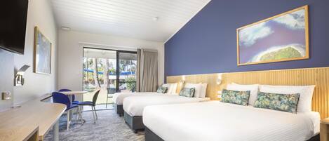 Habitación de hotel con dos camas, un escritorio y una silla. Se ve una ventana grande con vistas a palmeras y a la playa.