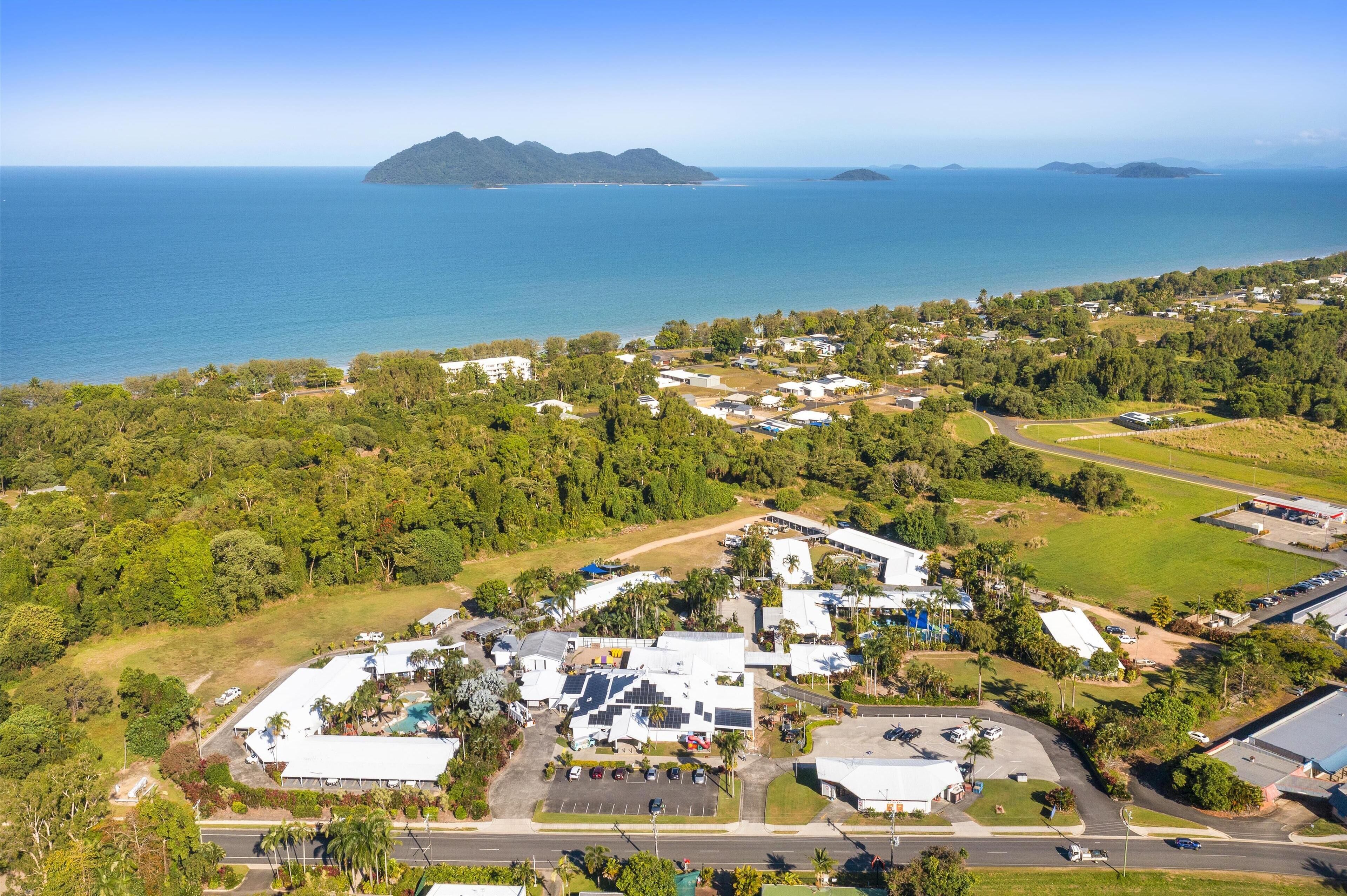An aerial view of a coastal area with a mix of residential buildings and greenery, a parking lot, and a road leading to the beach.