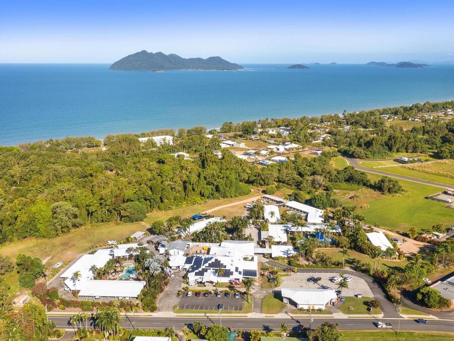 An aerial view of a coastal area with a mix of residential buildings and greenery, a parking lot, and a road leading to the beach.