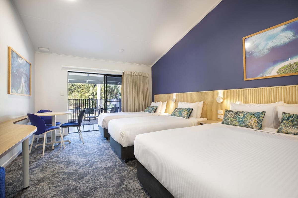 A hotel room with two beds, a desk, chairs, a large window, and a painting on the wall.