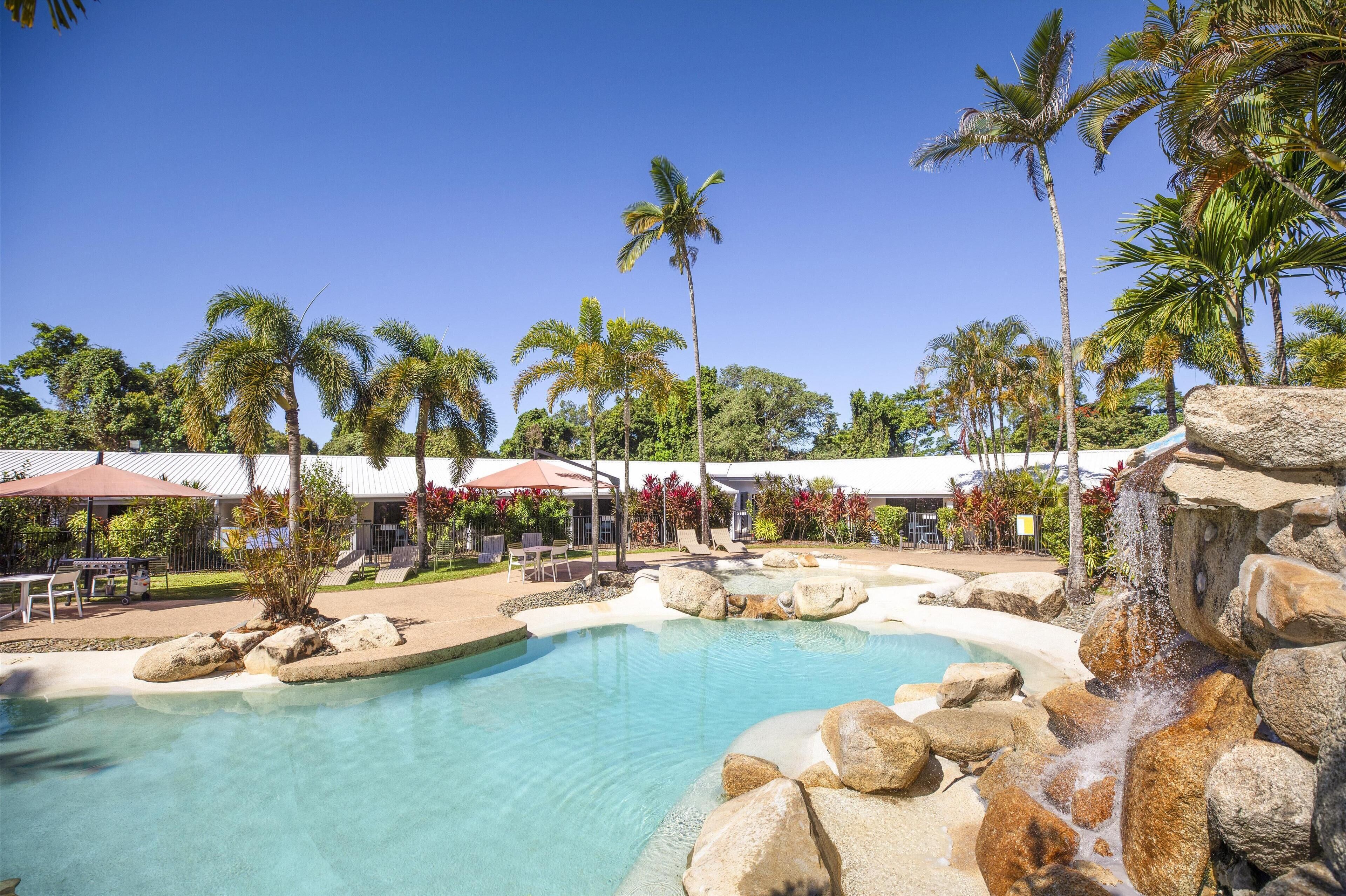 A pool area with clear water, surrounded by rocks and palm trees, and a building with a white roof in the background.