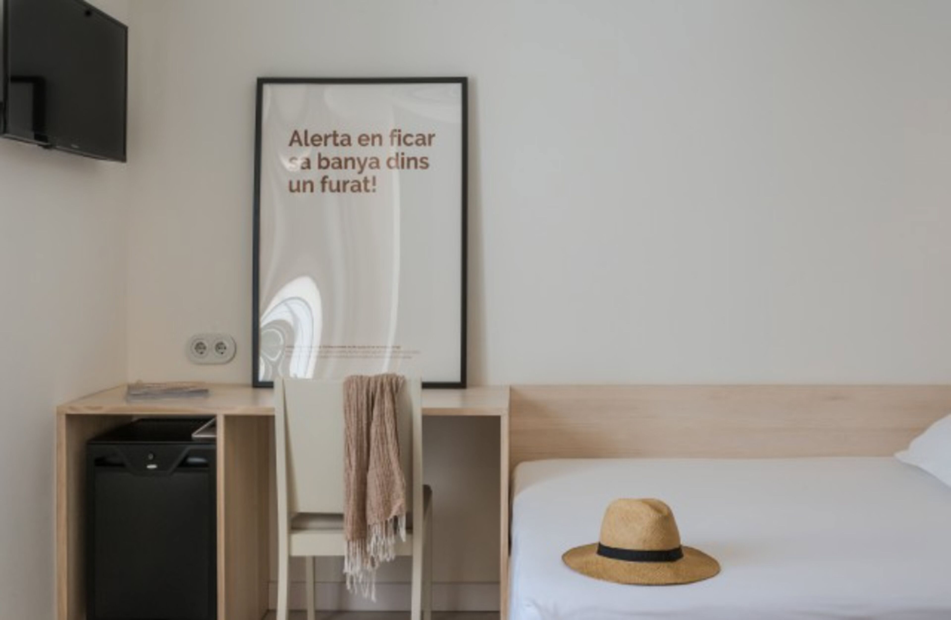 A hotel room with a bed, a desk with a chair, a television mounted on the wall, and a framed poster on the wall.