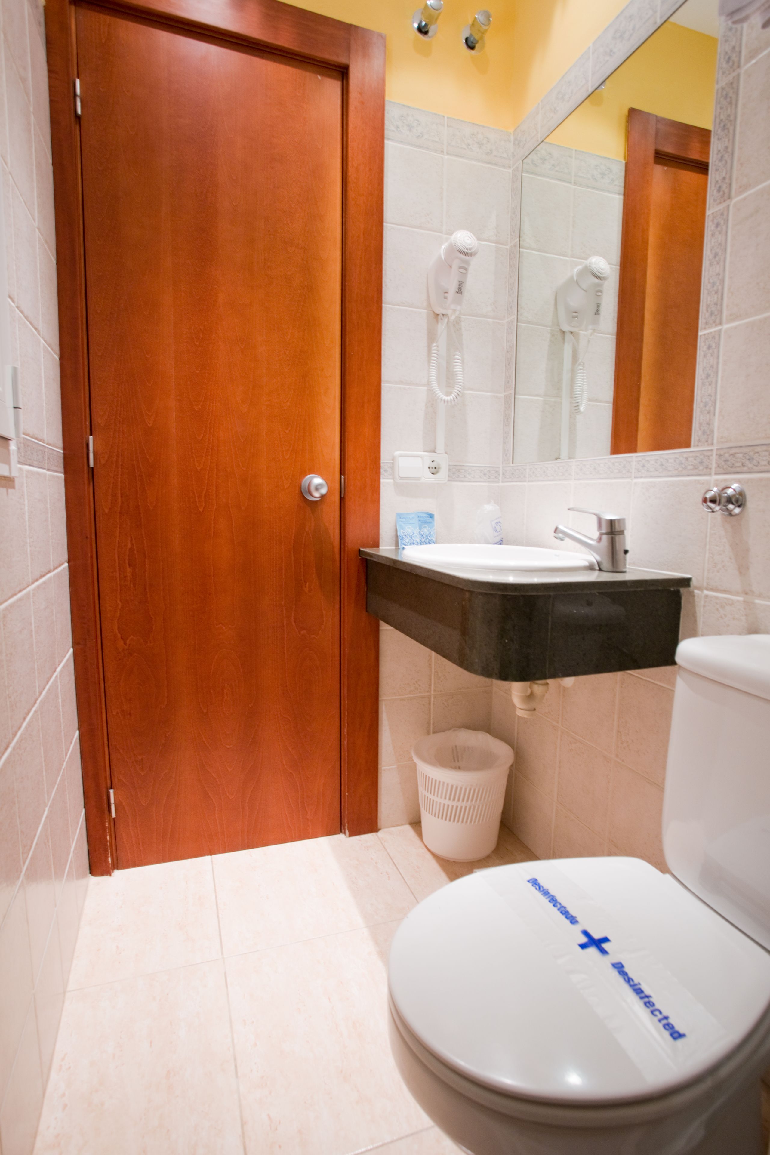 Un baño con puerta de madera, inodoro blanco, lavabo con encimera negra y espejo sobre el lavabo.