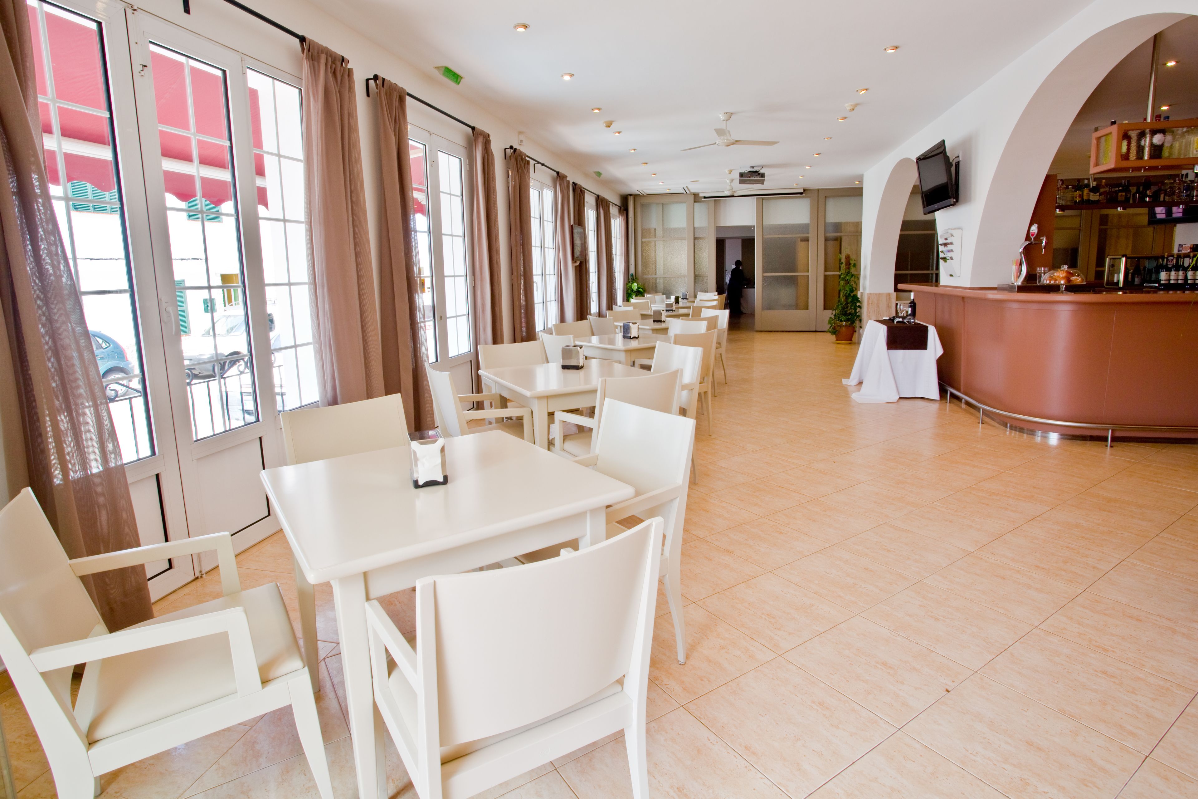 Un restaurante espacioso con mesas y sillas blancas, una zona de bar y amplias ventanas con cortinas.