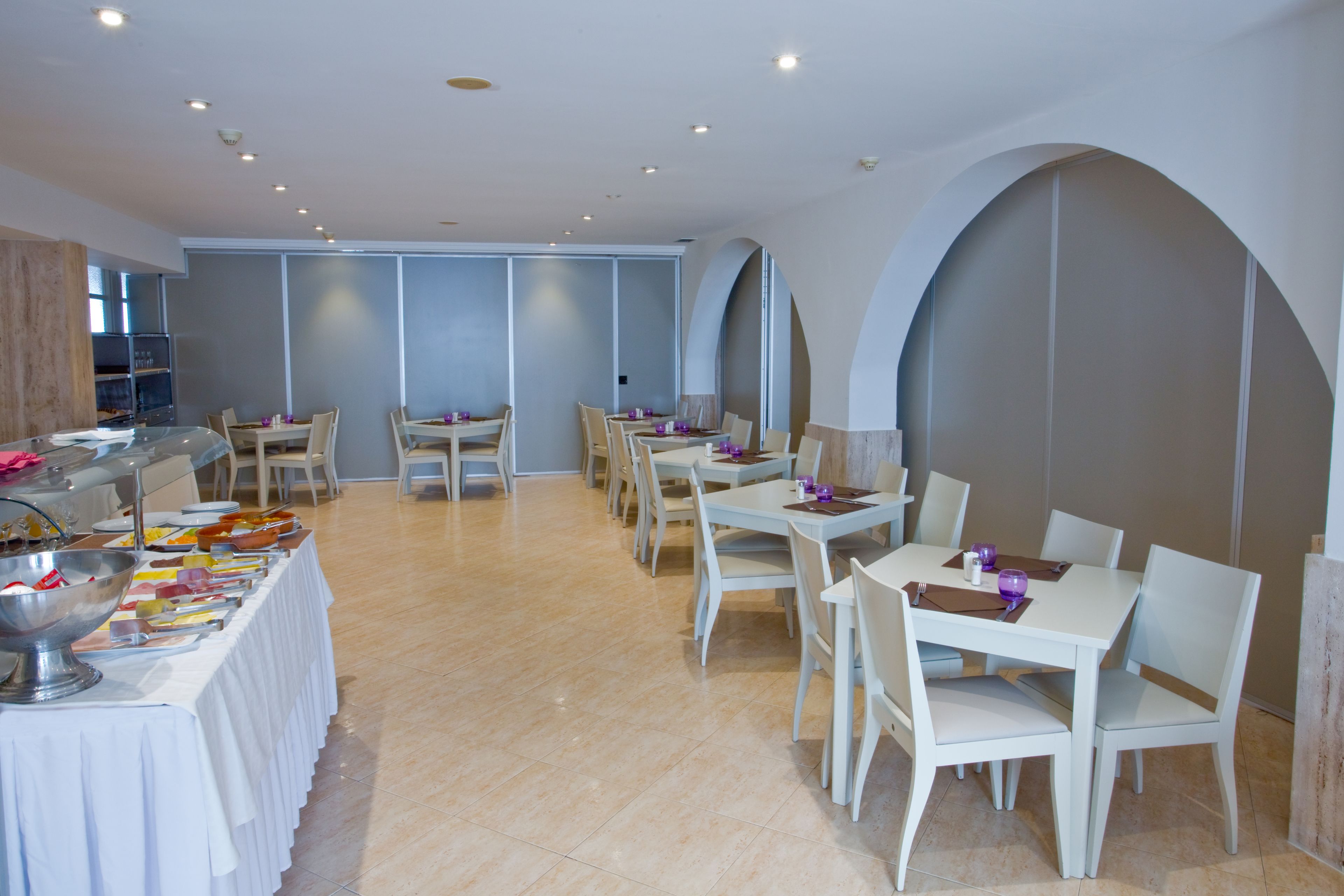 Un comedor con mesas y sillas blancas, una barra de buffet con diversos alimentos y un diseño interior moderno con techos abovedados e iluminación empotrada.
