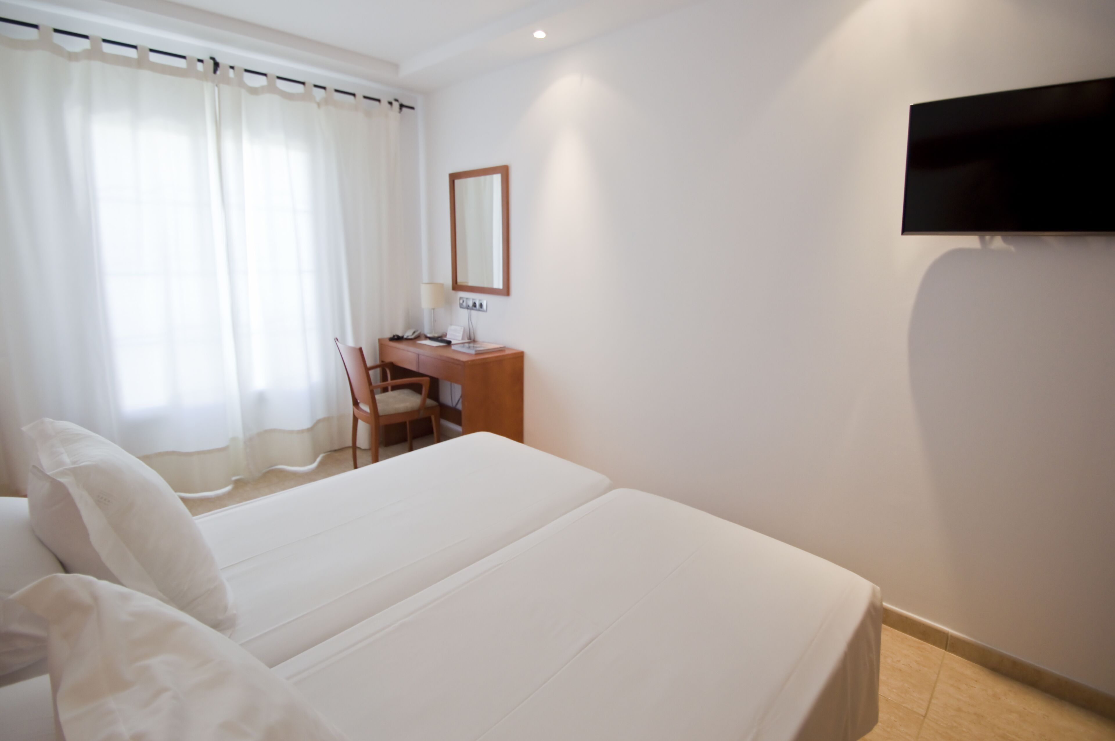 A hotel room with a large bed, a desk with a chair, a flat-screen TV mounted on the wall, and a window with sheer curtains.