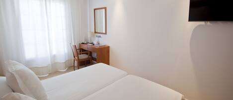 A hotel room with a large bed, a desk with a chair, a flat-screen TV mounted on the wall, and a window with sheer curtains.