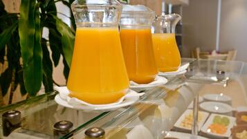 Une table de buffet avec des carafes en verre transparent remplies de jus d’orange, des assiettes blanches et divers aliments.