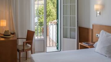 A hotel room with a bed, a chair, a desk with a lamp, and a view through an open door to a balcony.
