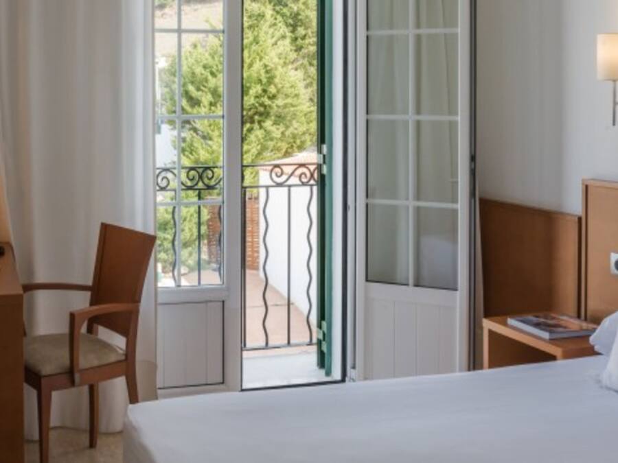 A hotel room with a bed, a chair, a desk with a lamp, and a view through an open door to a balcony.
