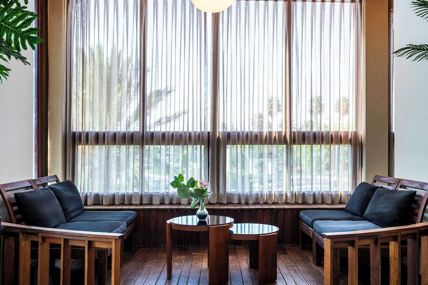 A room with two wooden chairs and a small round table, a vase with flowers, and a window with vertical blinds.