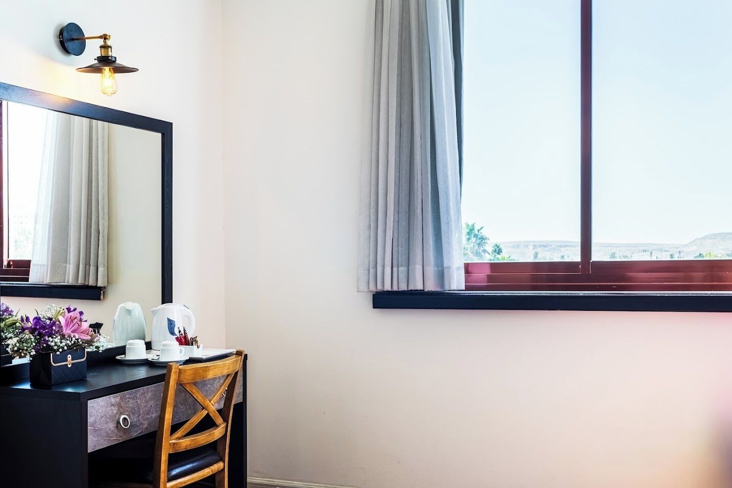 A hotel room with a large window, a wooden chair, a desk with a mirror, and a wall-mounted light fixture.