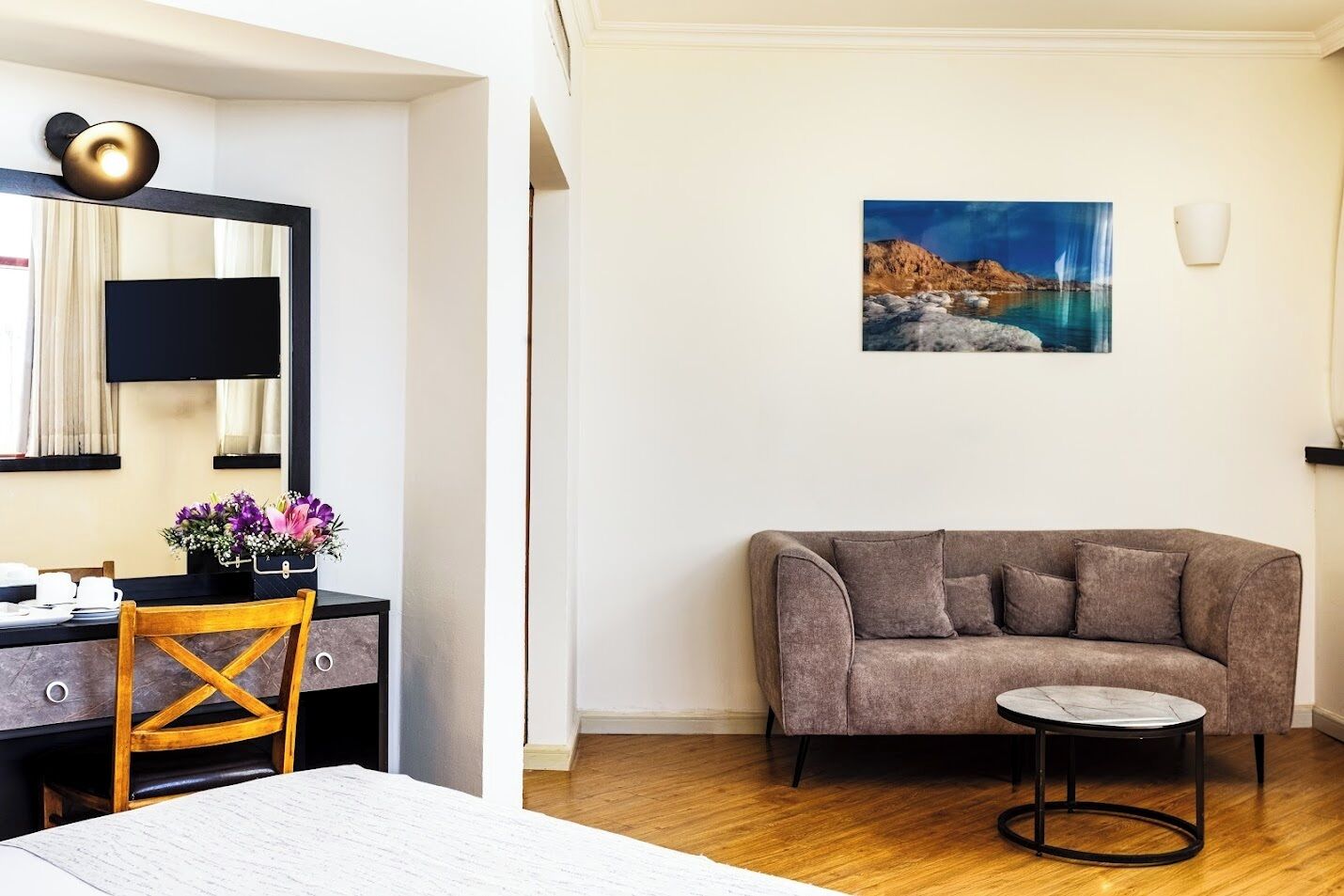 A modern living room with a sofa, a round coffee table, a wooden chair, a desk with a computer, and a painting of a coastal landscape.