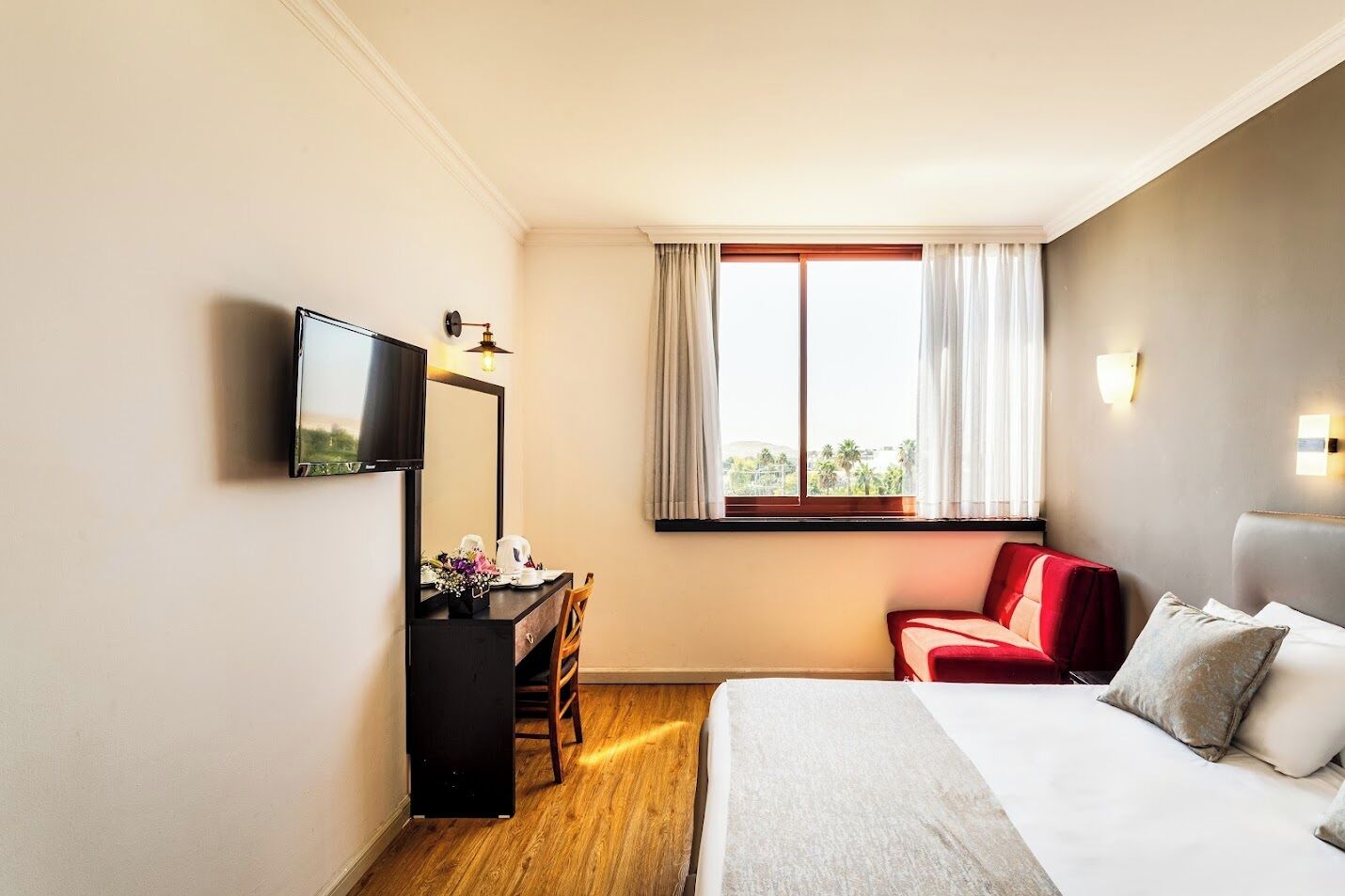 A hotel room with a bed, a desk with a chair, a TV, a window with curtains, and a wall-mounted light.