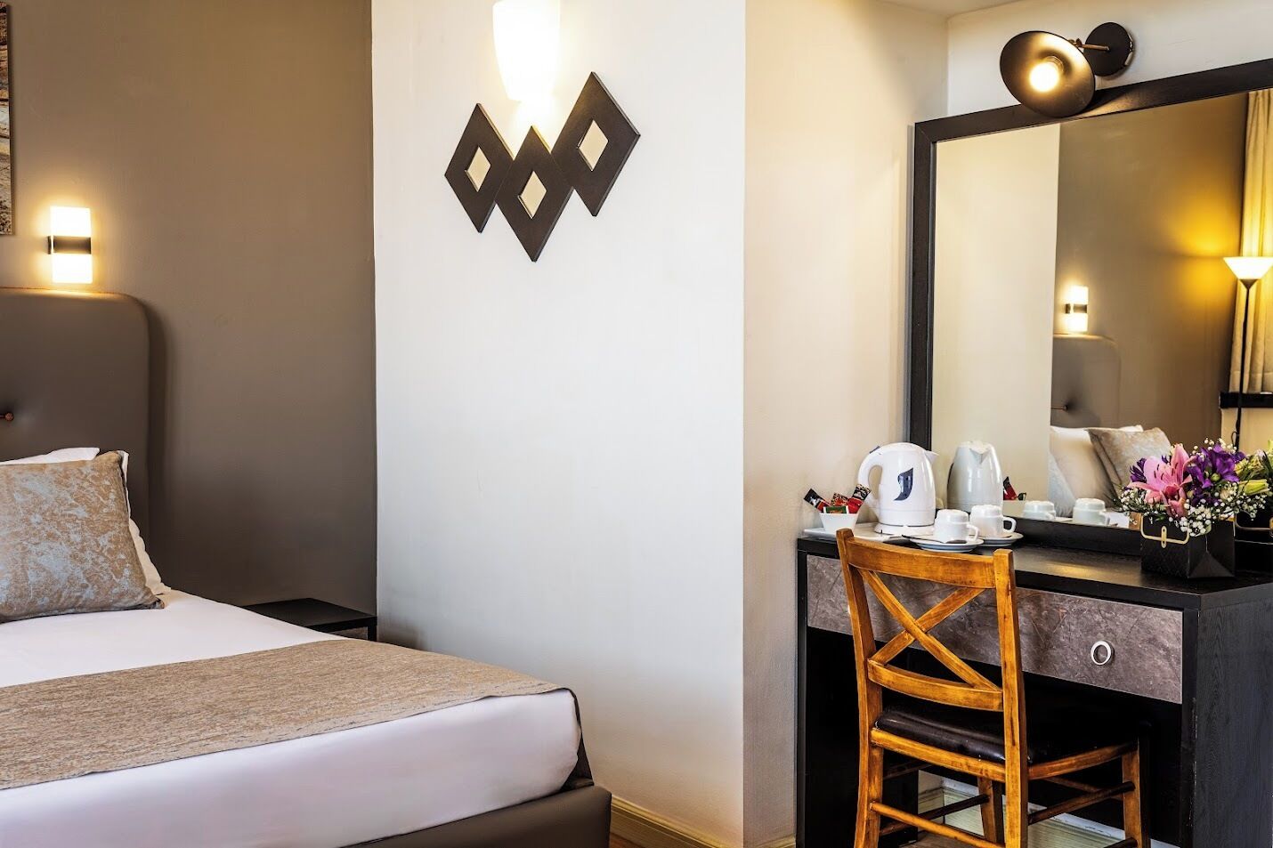 A hotel room with a bed, a desk with a chair, a mirror, and a wall-mounted light fixture.