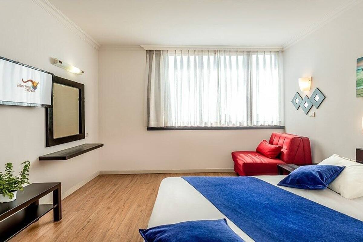 A hotel room with a bed, a red chair, a TV, and a window with curtains.