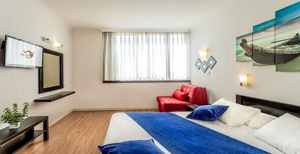 A hotel room with a bed, a red chair, a TV, and a window with curtains. - Inbar Hotel (Arad)