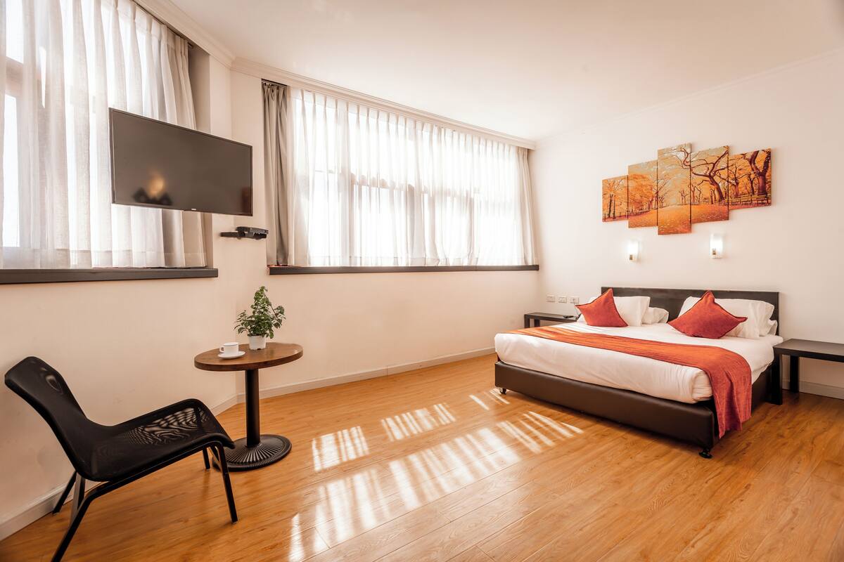 A modern hotel room with a bed, a TV, a small table with a plant, and a chair.