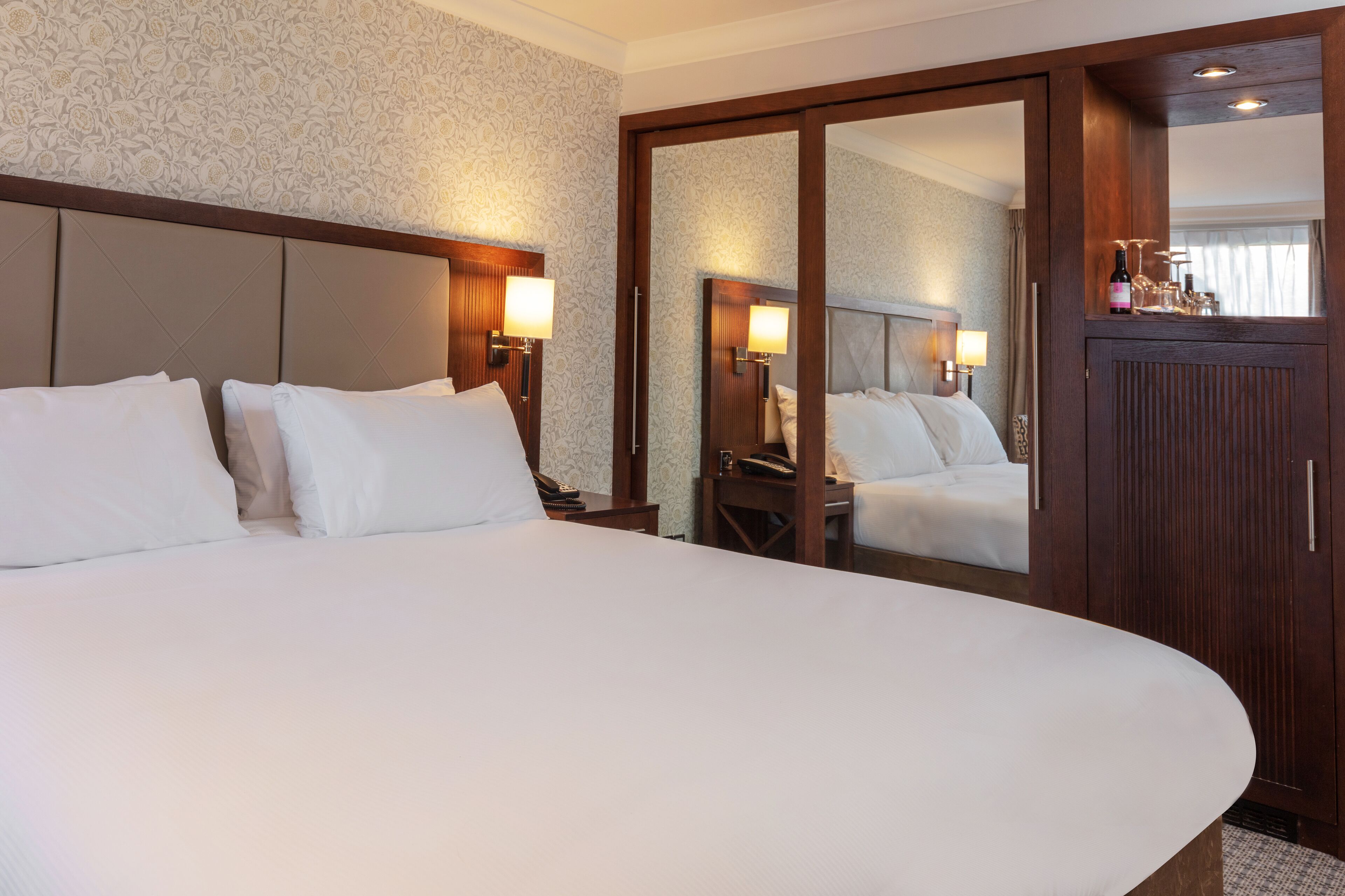 A large bed with white linens, two bedside tables with lamps, a wooden wardrobe, and a mirror reflecting the room.