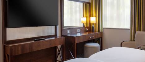 A hotel room with a large flat-screen TV, a desk with a mirror, two bedside lamps, a chair, and a bed.
