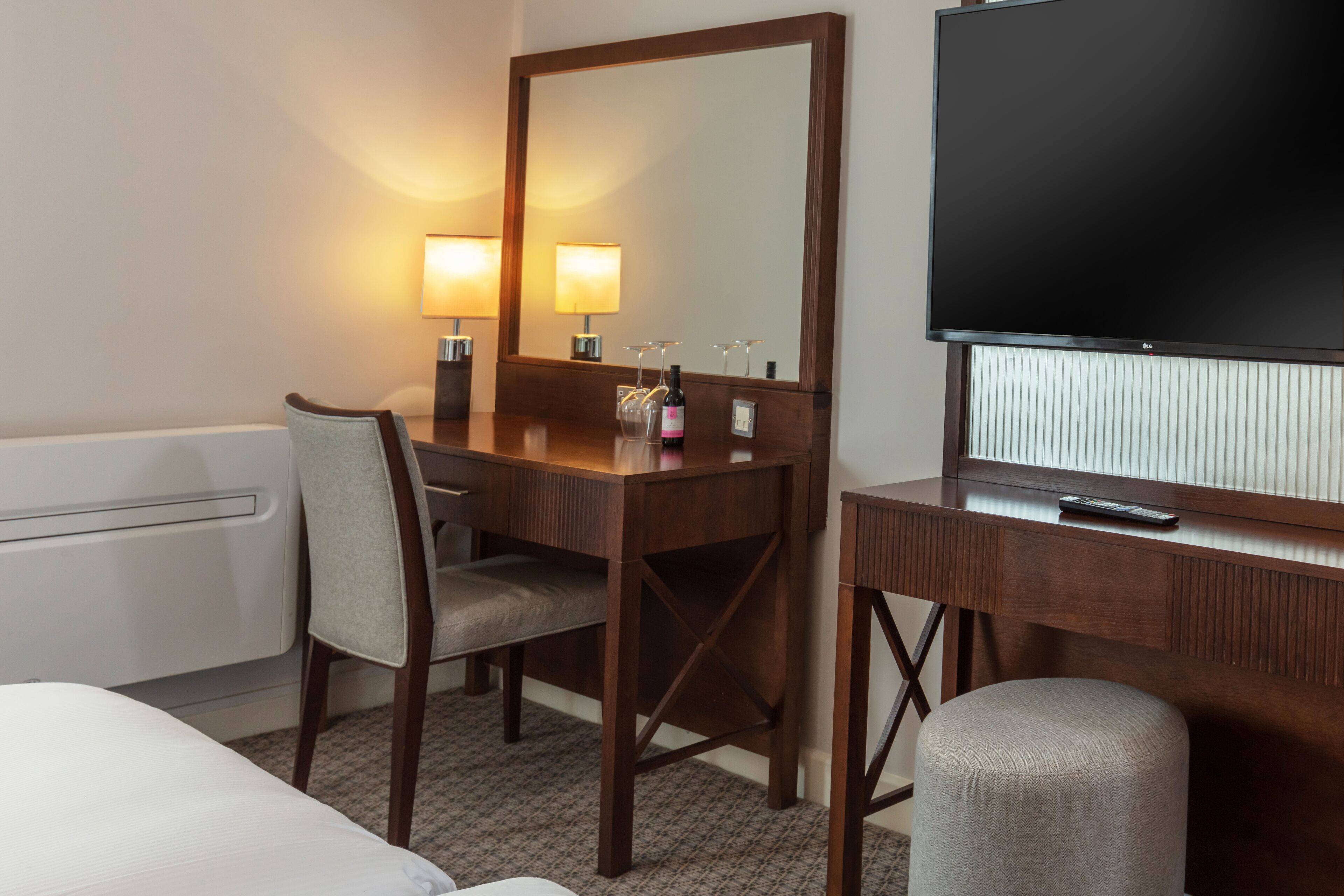A hotel room with a wooden desk, a large mirror, a television, a bed, and a chair.