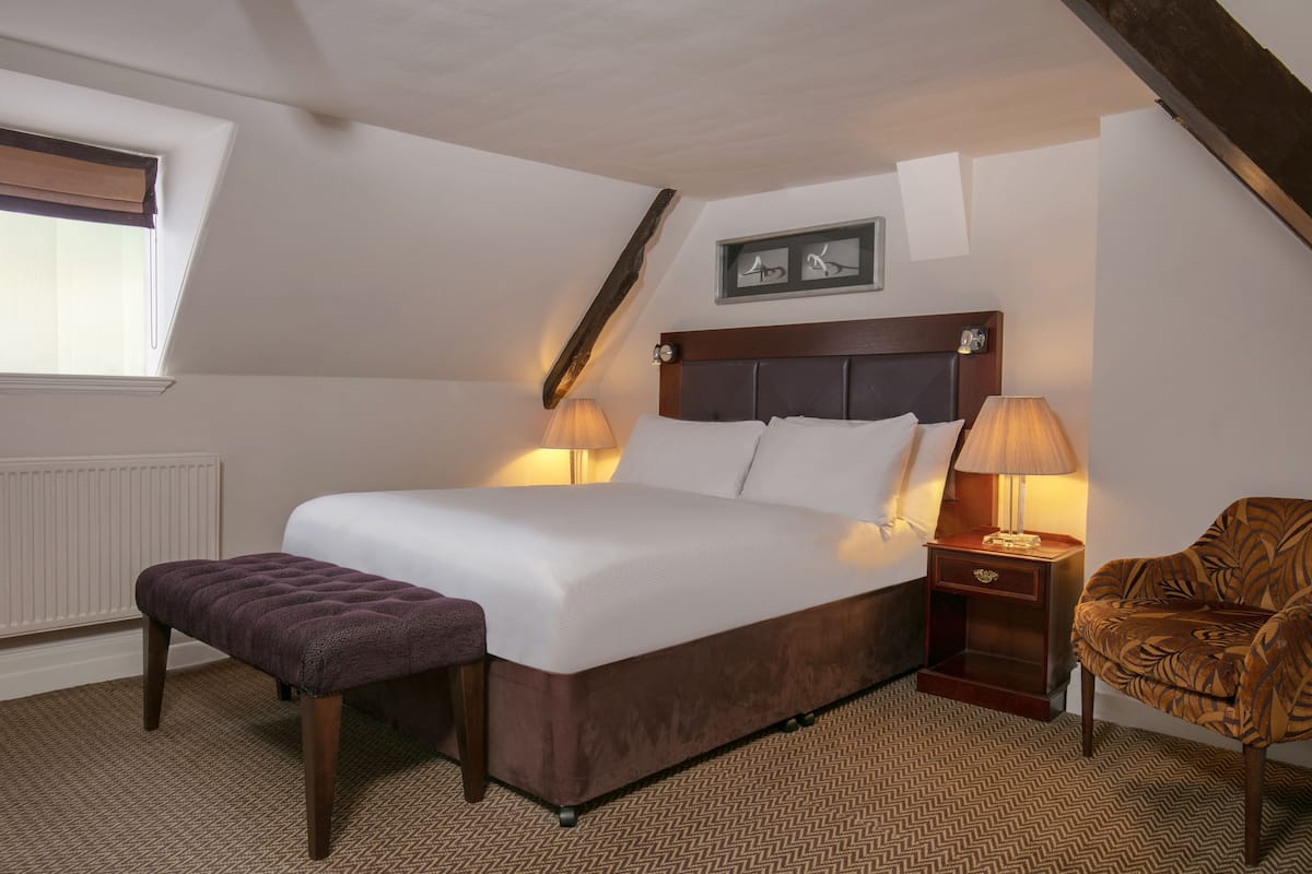 A neatly arranged bedroom with a large bed, two bedside tables with lamps, a wooden bench, and a patterned armchair.