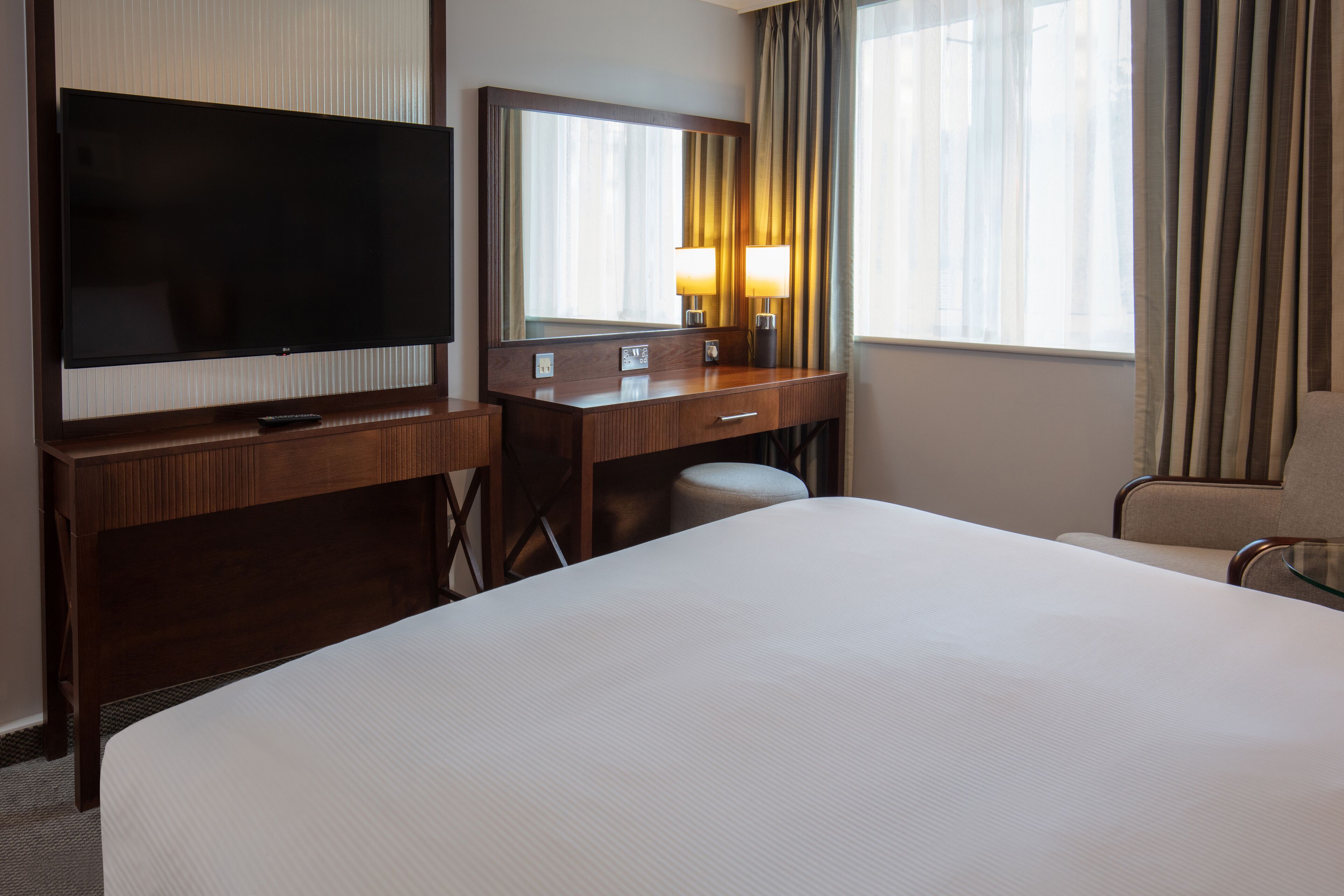 Una habitación de hotel con una cama grande, televisión, escritorio con espejo y una silla.
