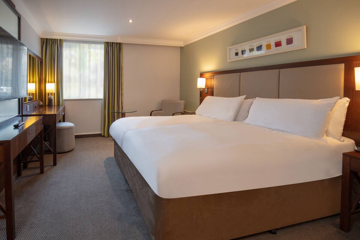 A hotel room with a large bed, a desk with a chair, a small stool, and a television.