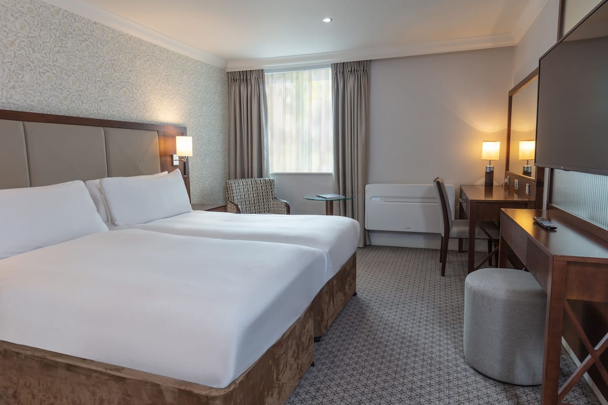 A hotel room with two beds, a desk, a chair, a small table, and a window with curtains.
