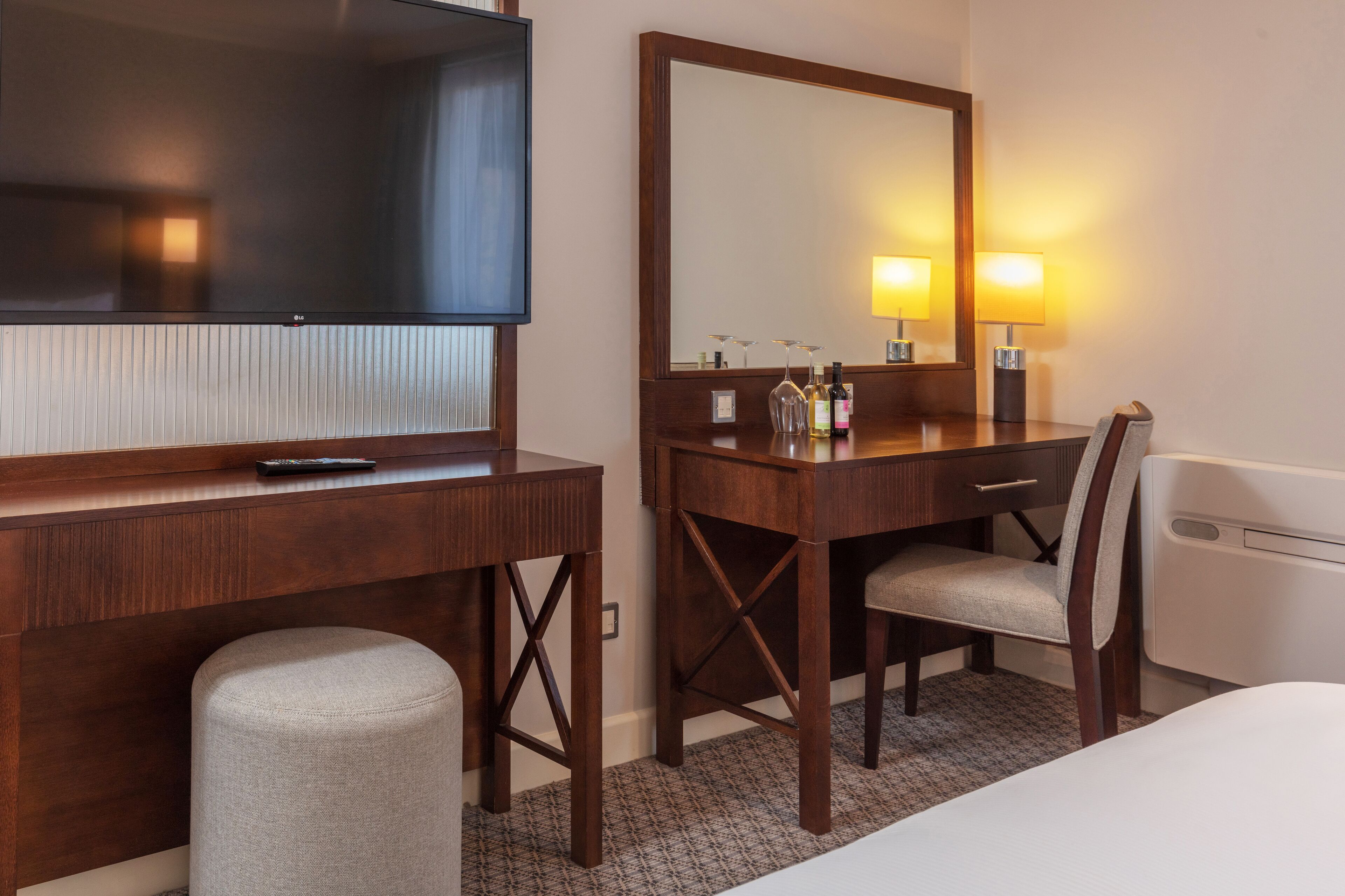 Habitación de hotel con escritorio de madera, un espejo grande, televisión, lámpara y una silla.