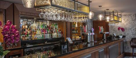 A bar area with a long counter, multiple shelves of liquor bottles, hanging wine glasses, and a patterned floor.