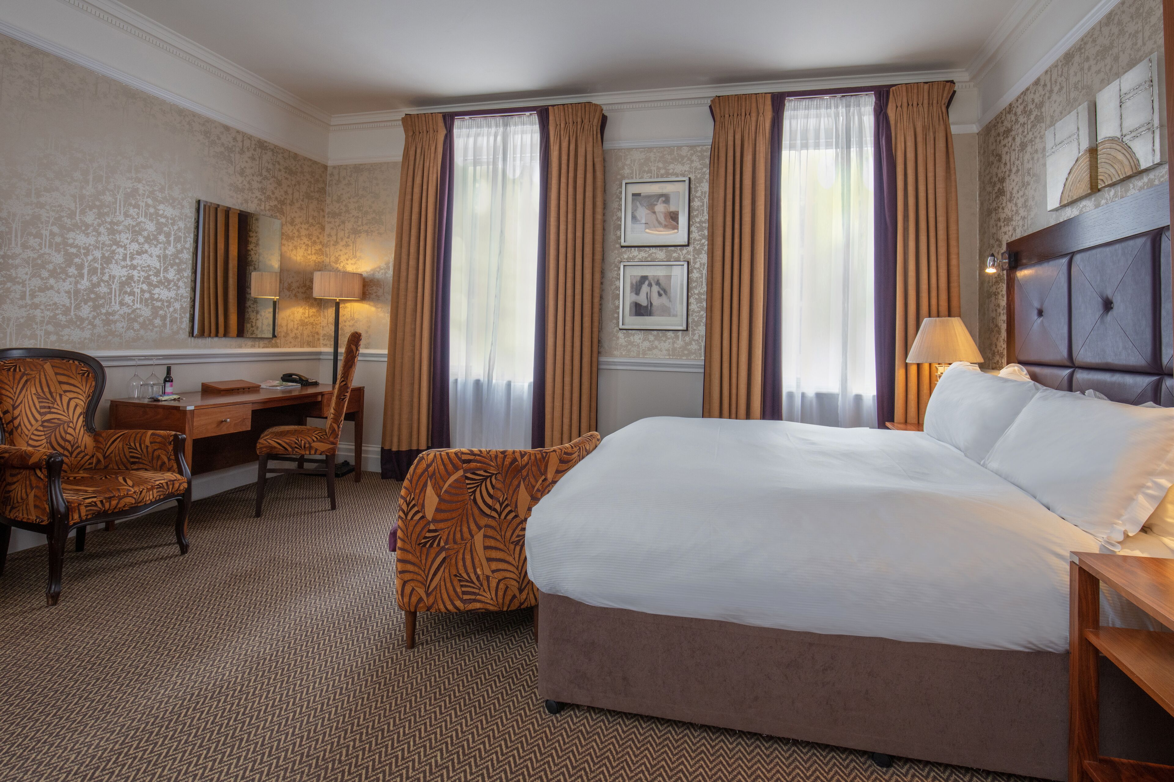A hotel room with a large bed, a desk with a chair, a lamp, and a picture on the wall.