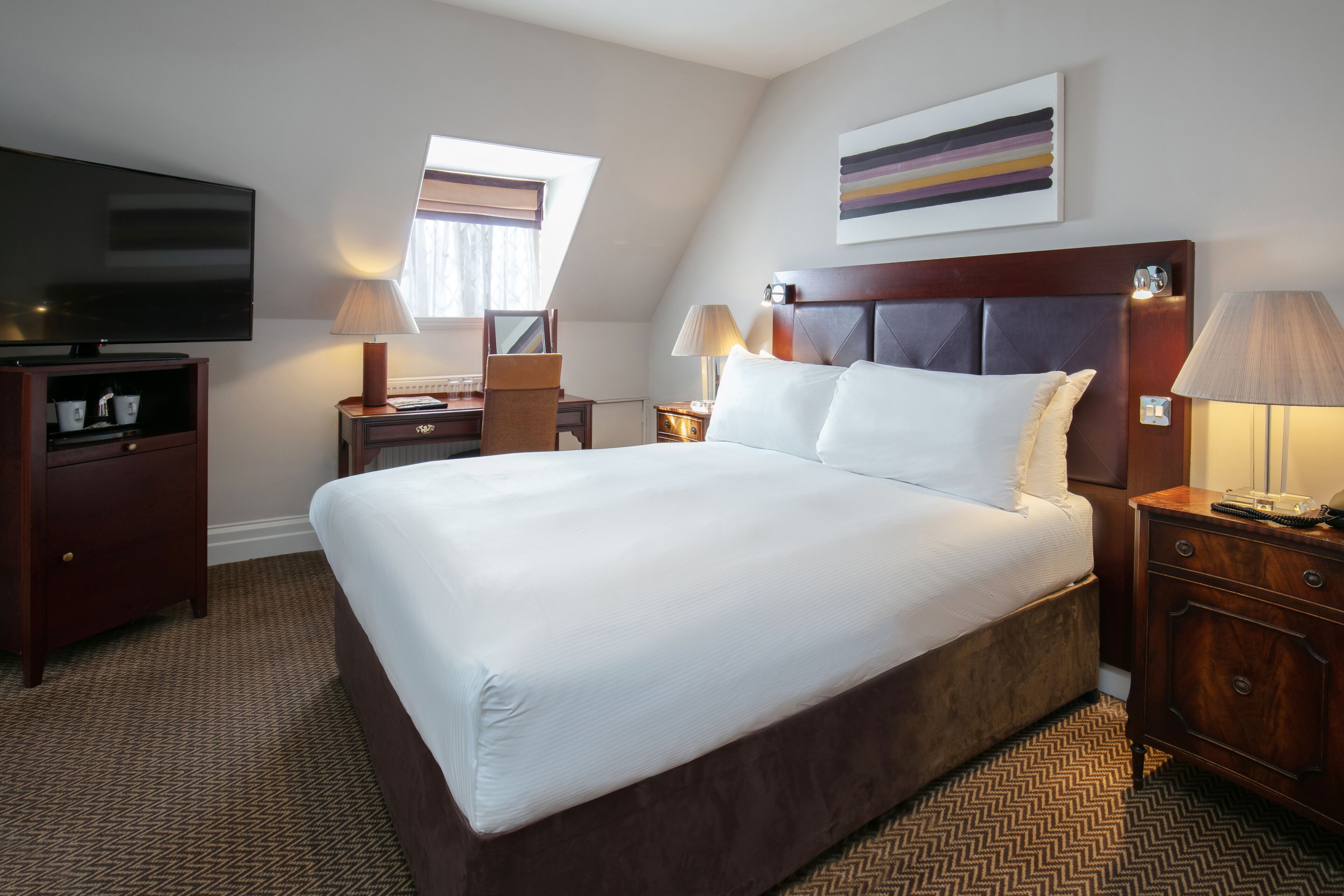A hotel room with a large bed, a flat-screen TV, a desk with a lamp, and a window providing natural light.