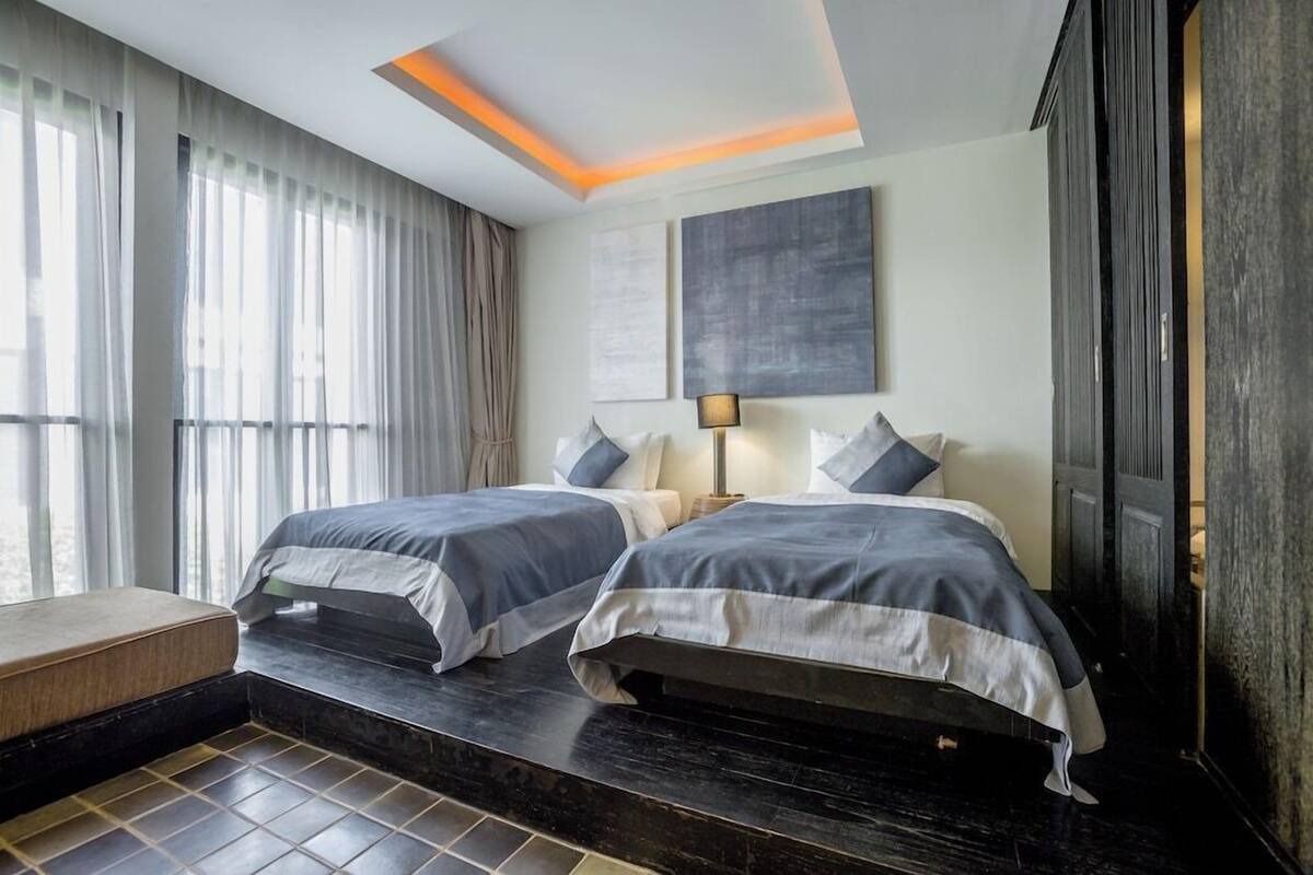 Habitación de hotel con dos camas, un sofá, un ventanal con cortinas, una lámpara en el techo y obras de arte en la pared.