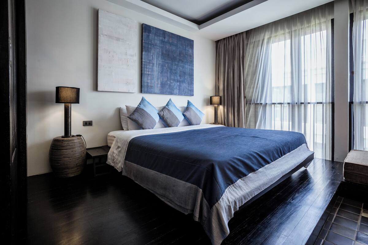Un dormitorio moderno con una cama grande, un cuadro abstracto en la pared y vistas al exterior a través de cortinas de gasa.