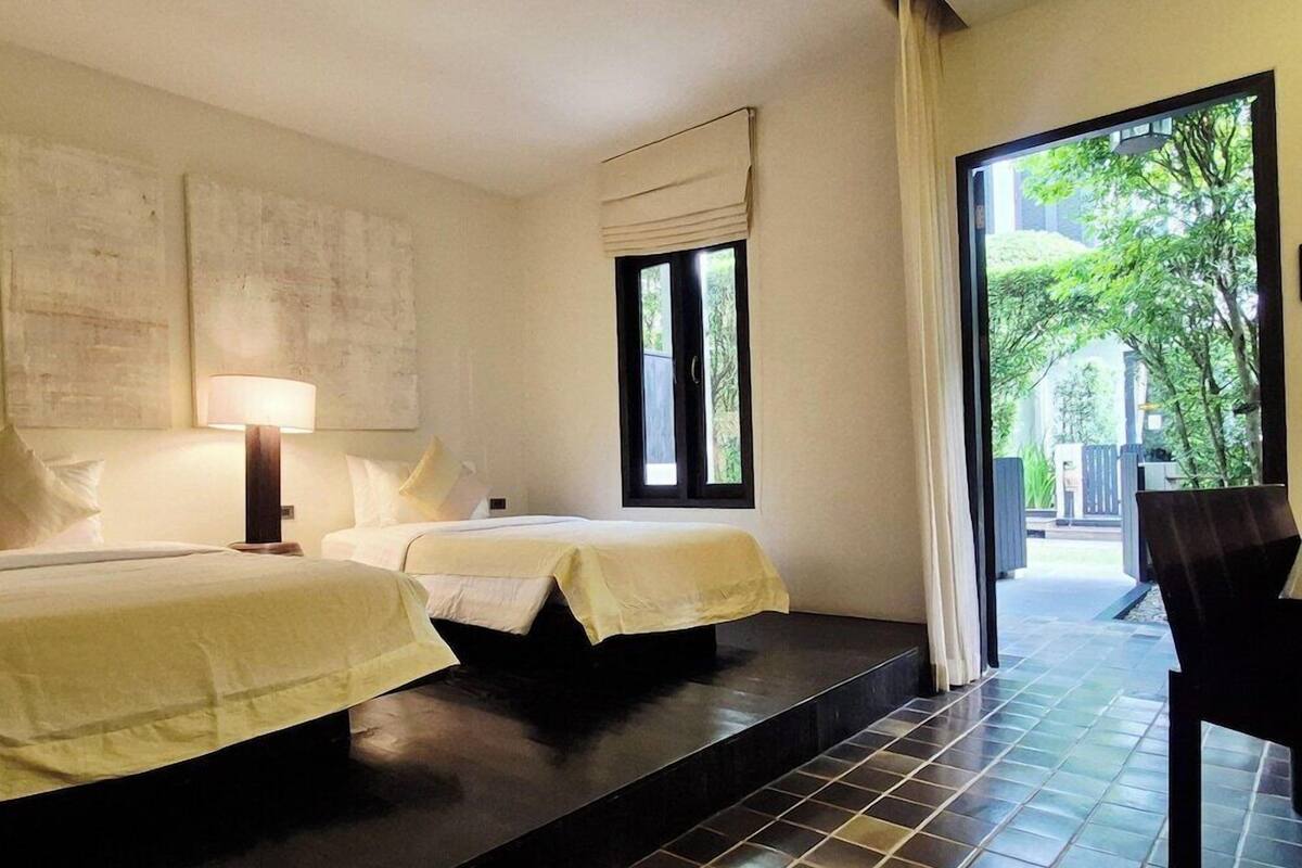 Habitación de hotel con dos camas, una mesita de noche, una lámpara, un ventanal y vistas a zonas verdes.