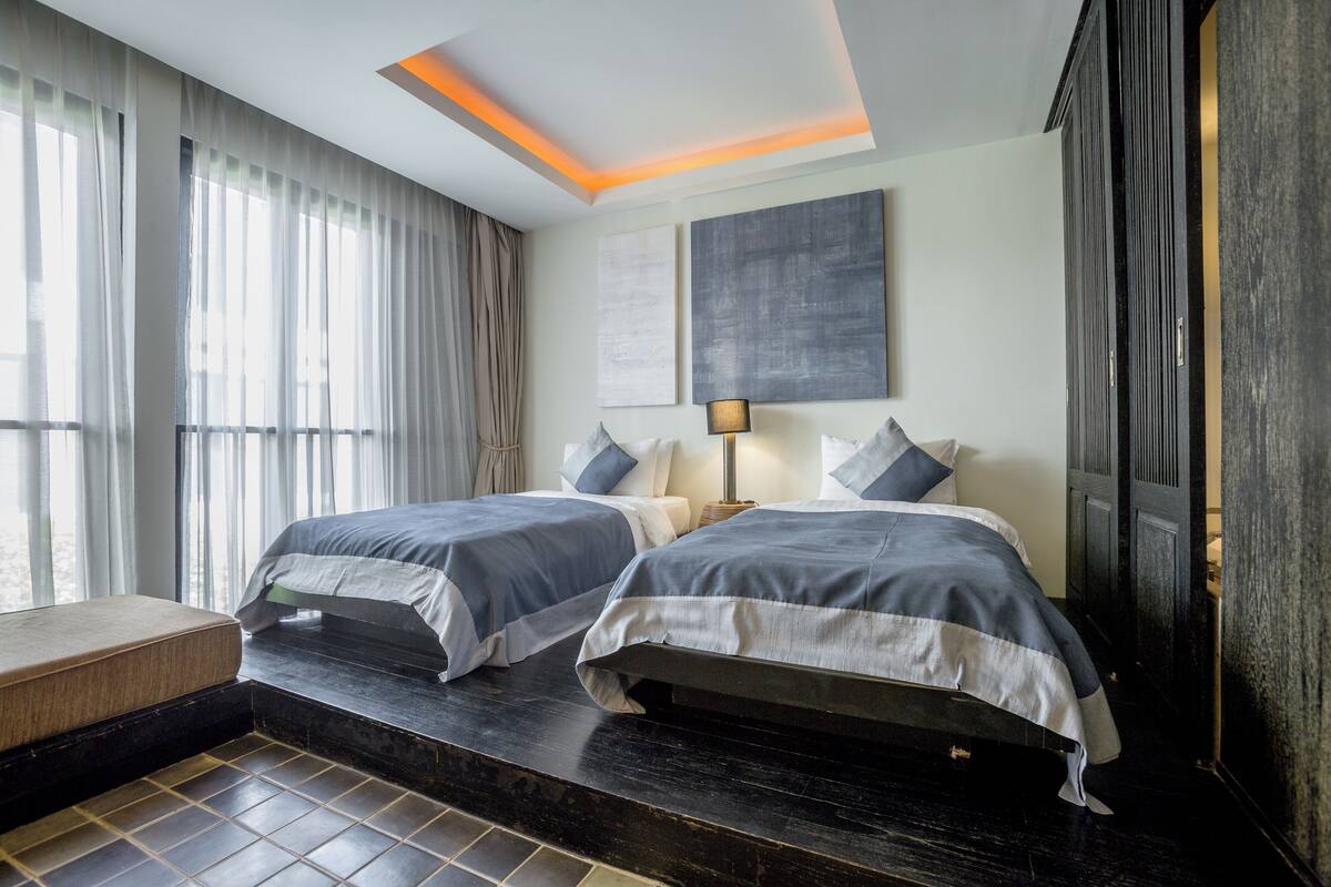 Una habitación de hotel moderna con dos camas, un ventanal con cortinas traslúcidas, una lámpara en el techo y obras de arte en la pared.
