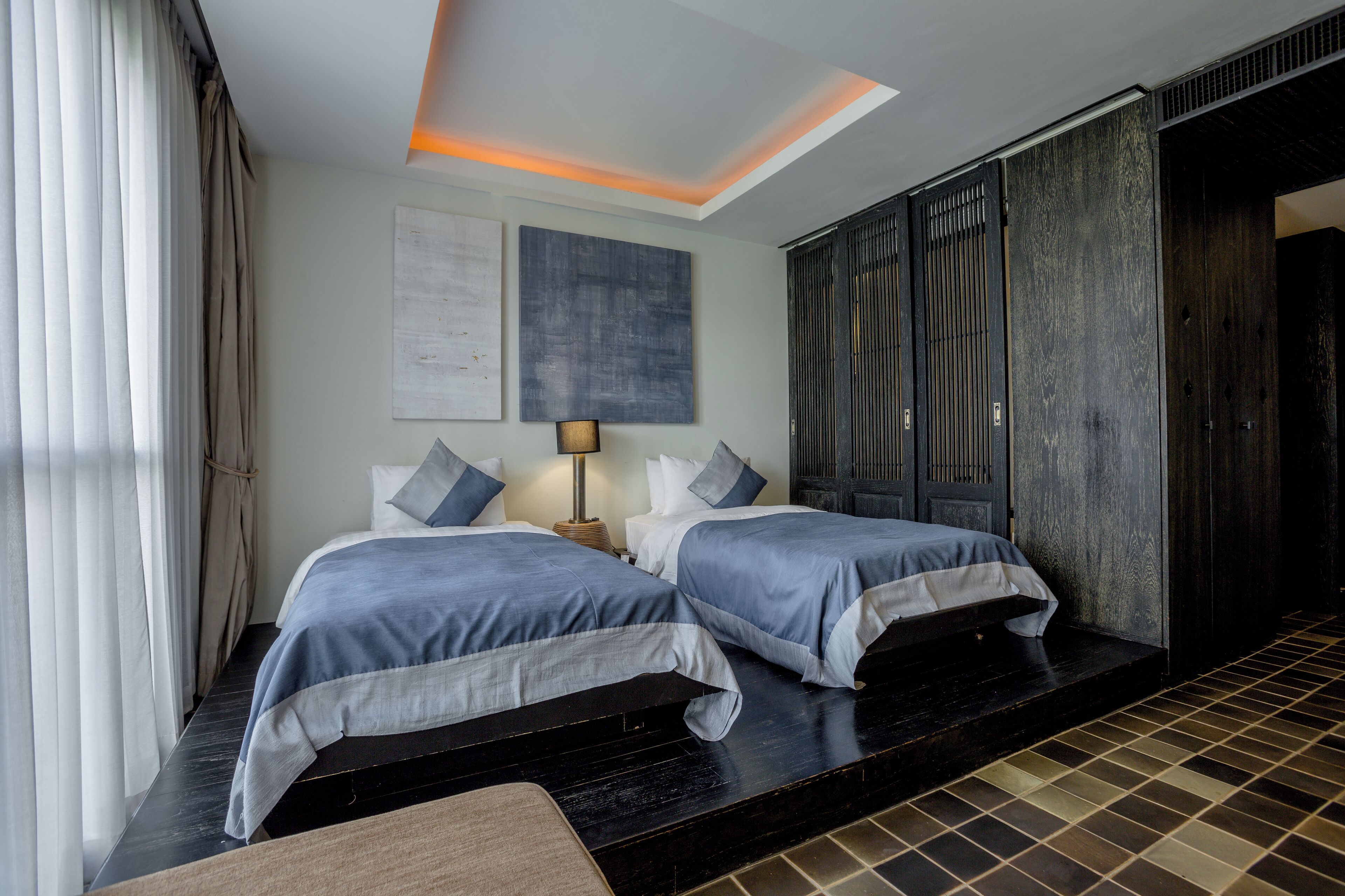A hotel room with two beds, a large dark wooden wardrobe, a ceiling light, and a view of the outside through the window.
