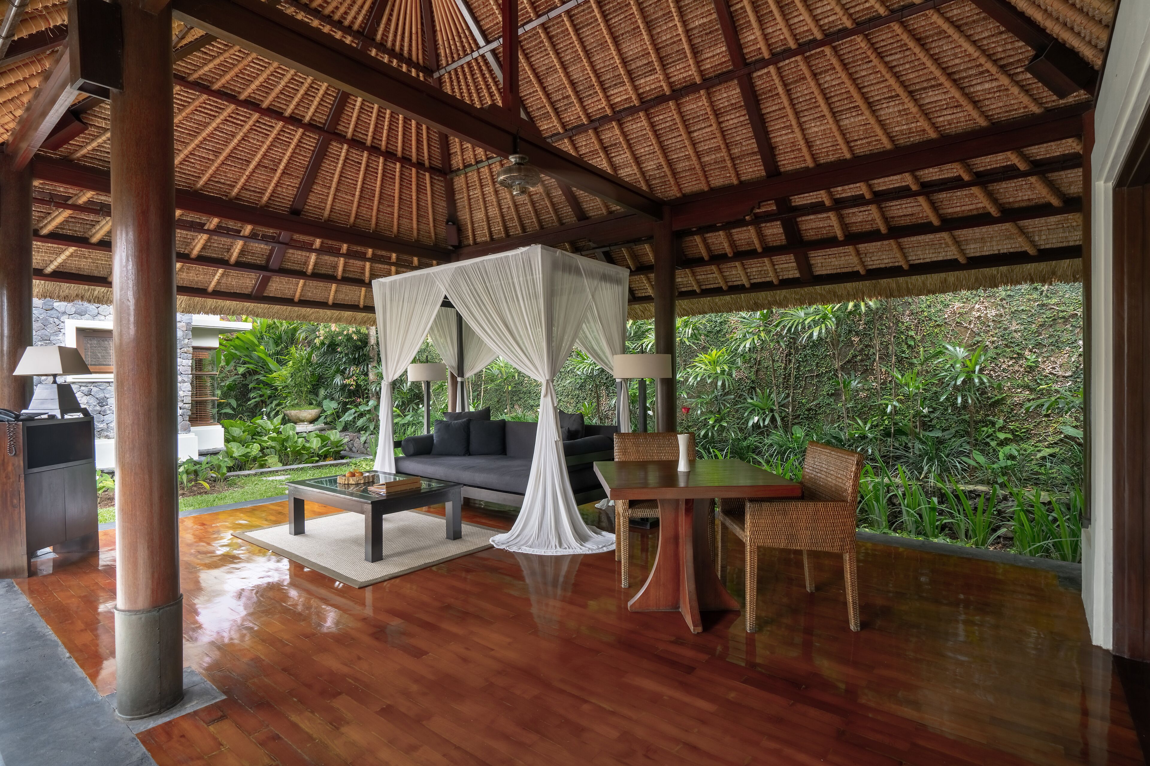 An open-air room with a wooden floor, a thatched roof, a sofa, a table, and chairs.