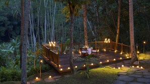 A wooden deck with a table and chairs, surrounded by trees and lit by candles. - Kayumanis Ubud Private Villas & Spa (Ubud)