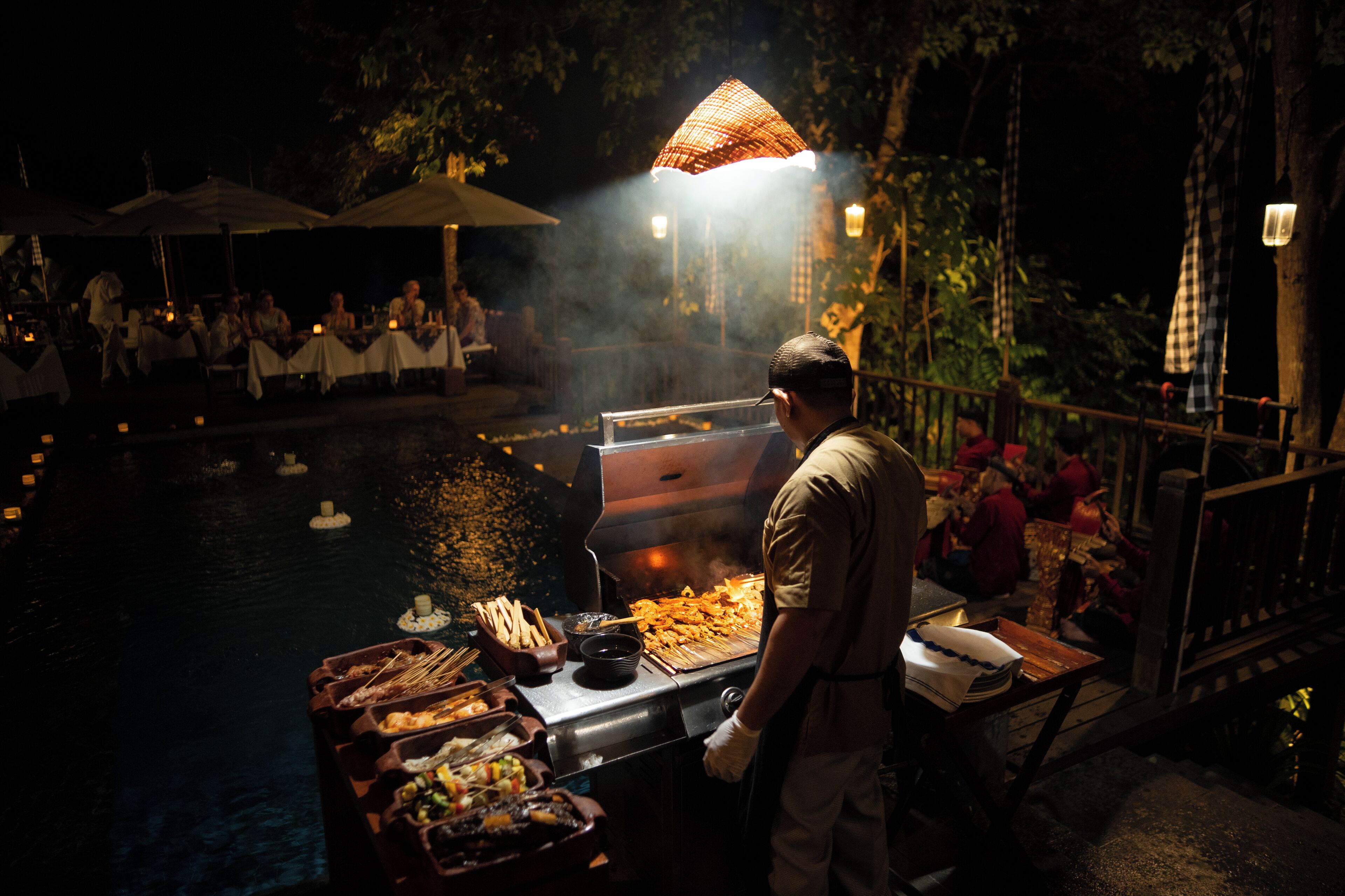 A nighttime outdoor dining area with a grill station, tables, and umbrellas.