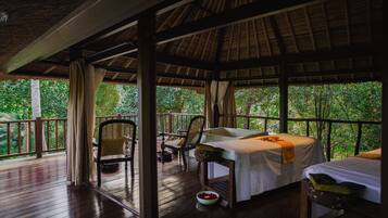 A wooden deck with a thatched roof, two beds, a chair, and a table.